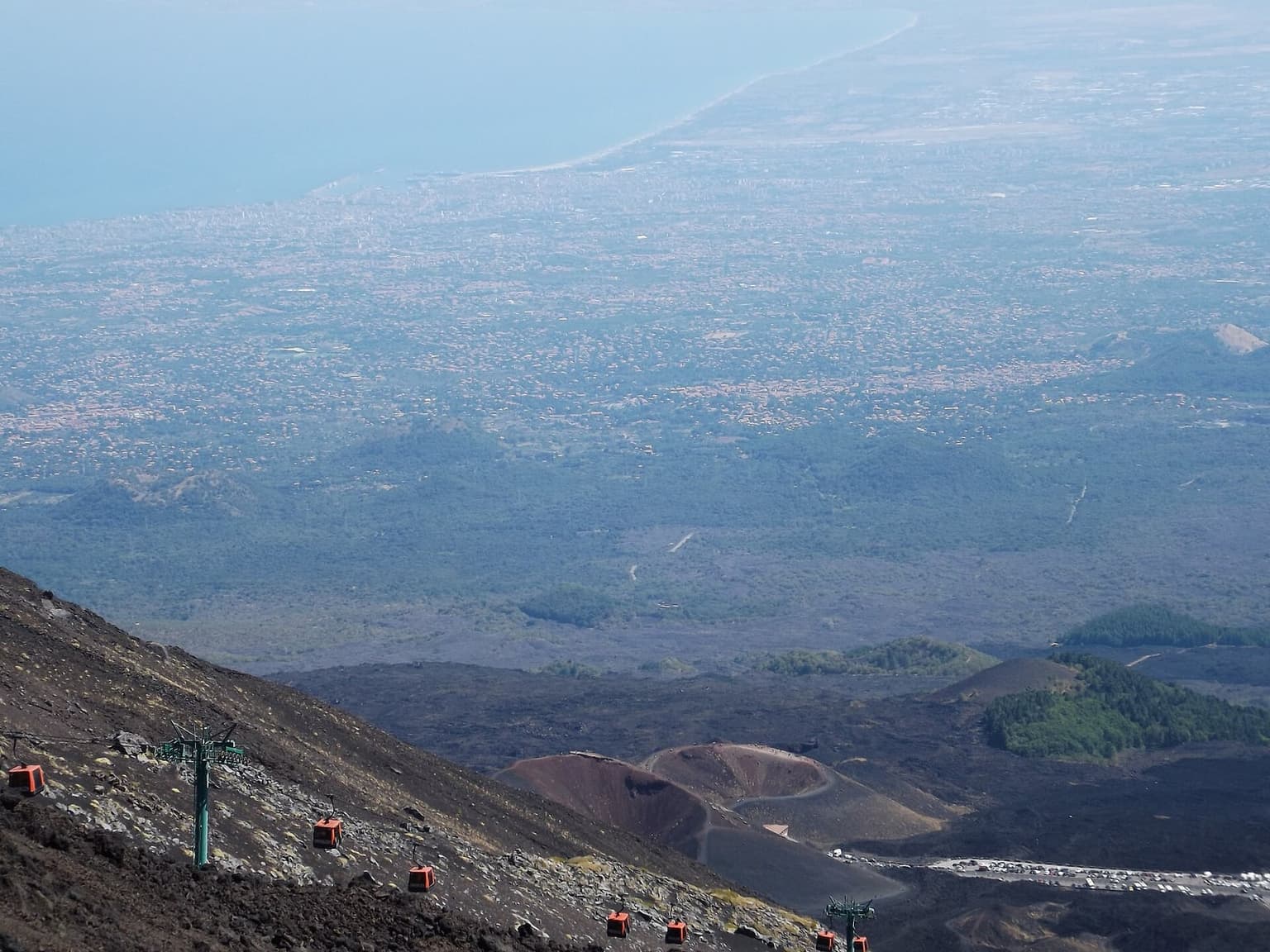 Etna cable-car gondolas descending a black lava slope, with a red volcanic cone, pine-covered hillside, and the hazy Catania coastal plain and sea visible in the distance.