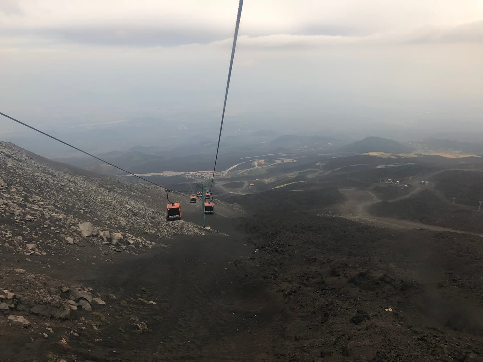 View from an Etna cable-car cabin looking along the ascending wire, black lava rock filling the foreground slope and a hazy coastal plain stretching to the sea far below.