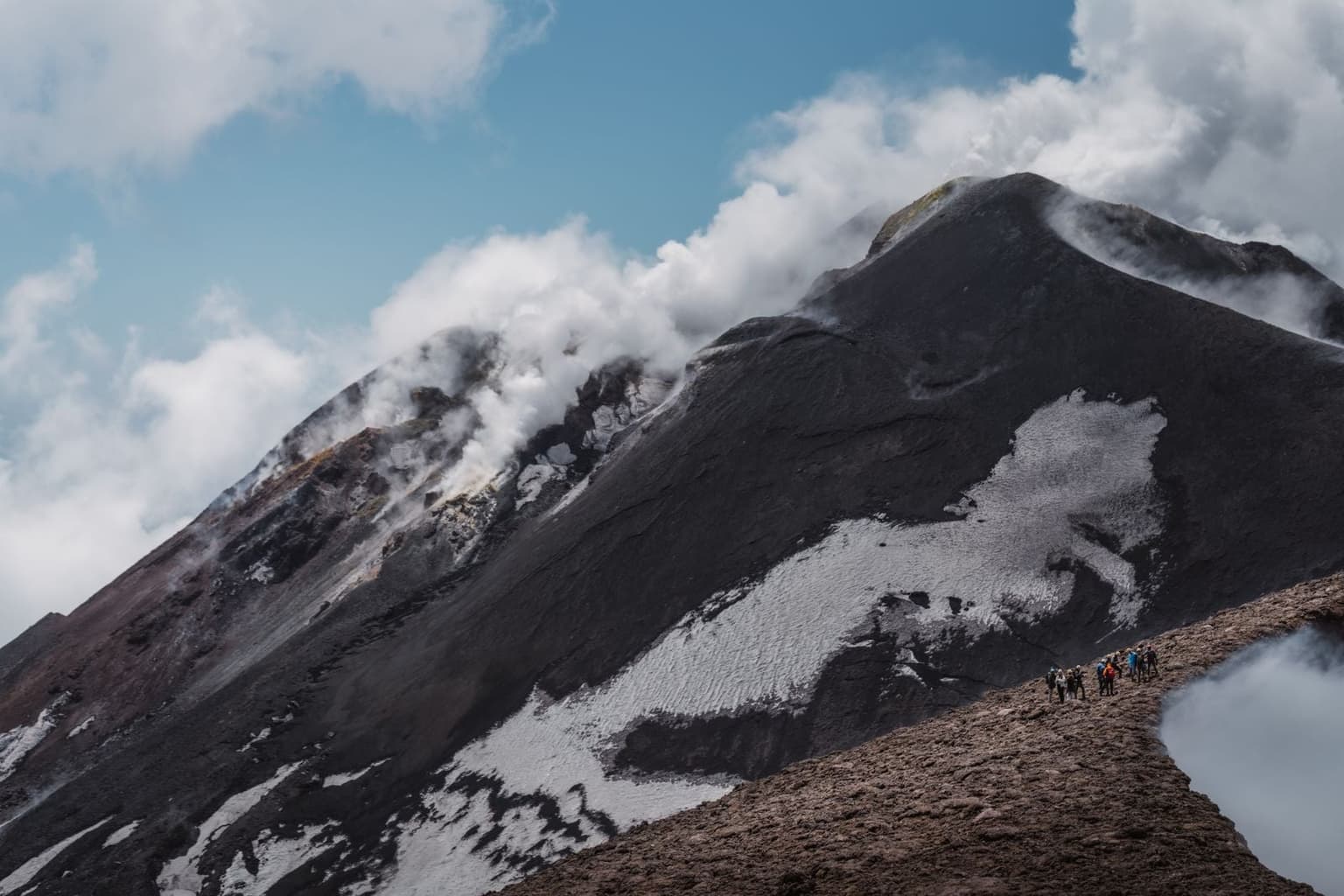 A line of hikers walks a narrow ridge path at the base of Mount Etna's summit cone as thick white steam vents directly from the crater rim above them, snow patches streaking the dark slopes.