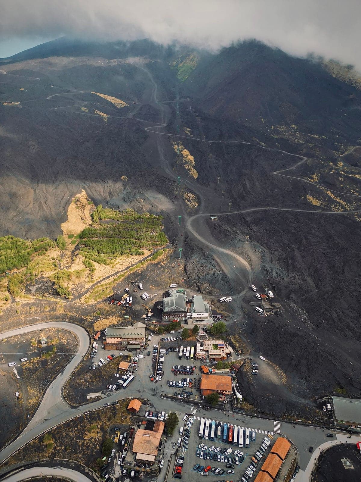 Aerial view of the Rifugio Sapienza base station on Mount Etna, its cable-car building and large parking lots packed with buses and cars, switchback roads climbing through black lava fields toward the cloud-wrapped summit above.