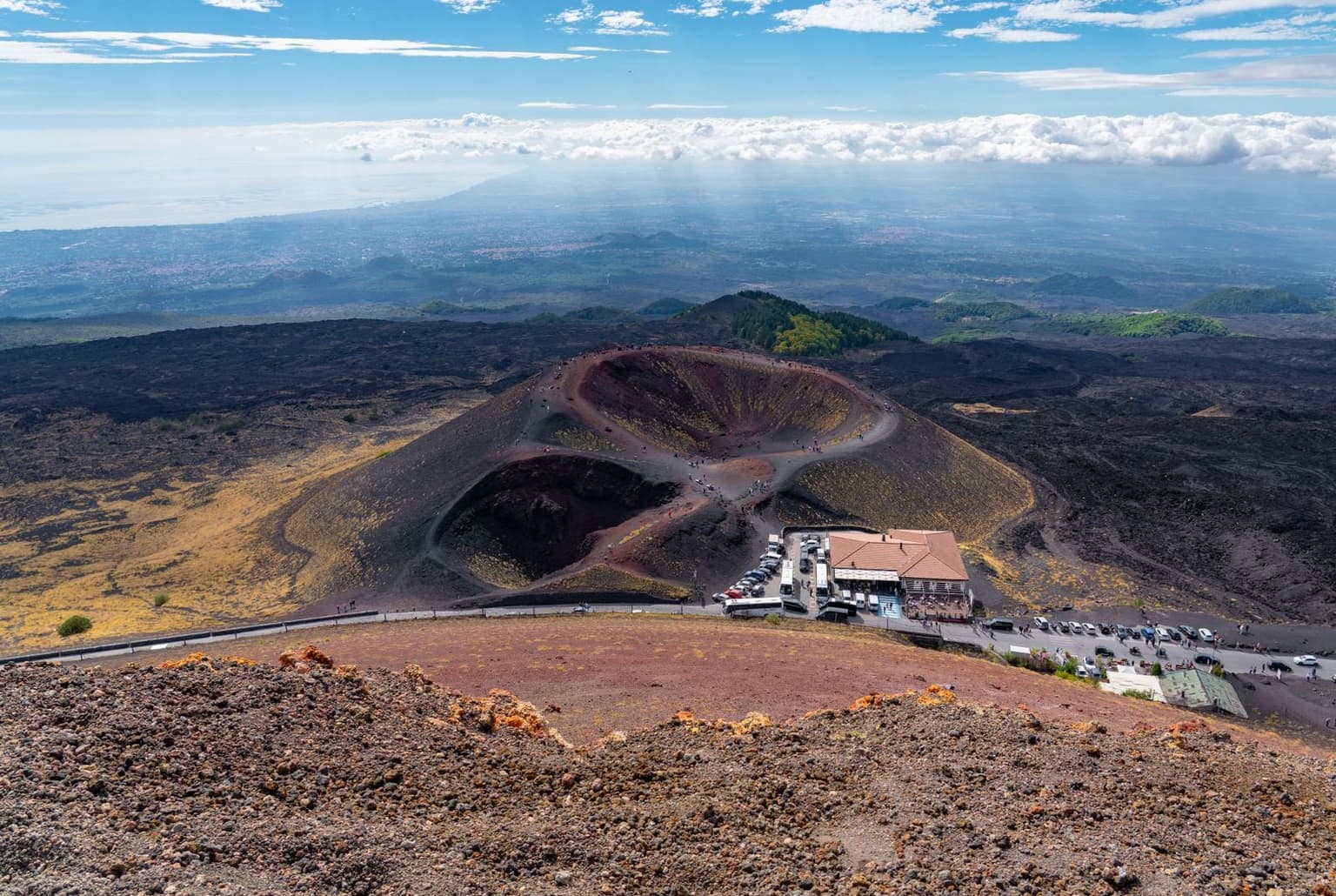 Aerial view of the twin red-and-black cinder Silvestri craters on Mount Etna, with visitors walking the rim path and a cluster of cars and tour buses parked at a roadside building below.