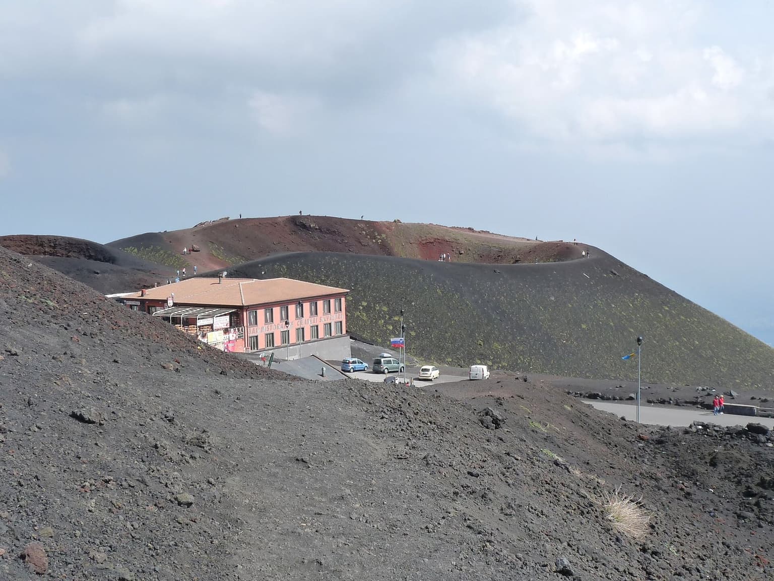The Silvestri crater cone with its rim path dotted with visitors, the pink Bar Ristorante Crateri Silvestri building and a small parking area with several cars at its base.