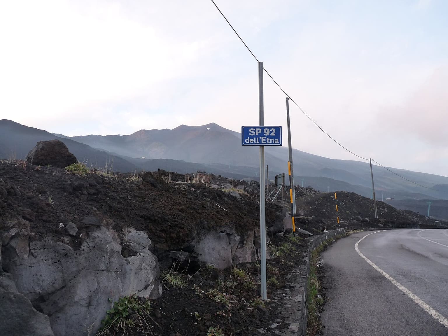 A blue road sign reading "SP 92 dell'Etna" beside the switchback road, with black lava fields on both sides and Etna's misty, smoking summit in the distance.