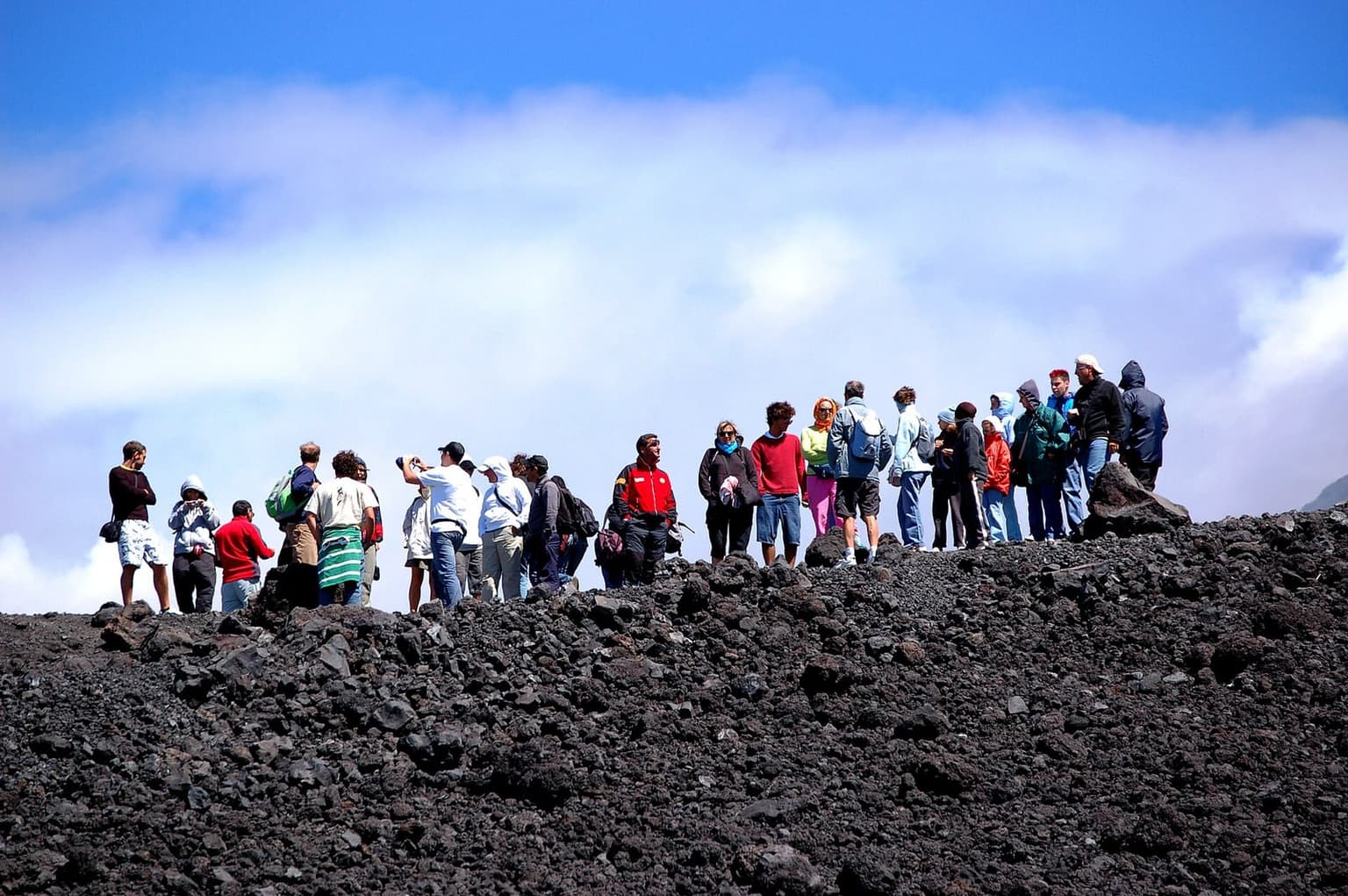 A large group of tourists in mixed casual jackets and backpacks standing together on a black volcanic ridge high on Etna, with cloud below and blue sky above.