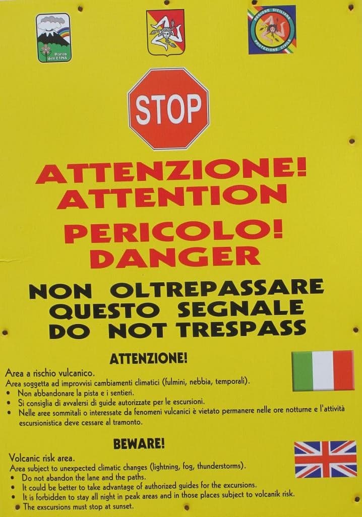 A yellow warning sign on Etna's upper slope, in Italian and English, marking a volcanic risk area and advising hikers not to go beyond it without an authorized guide.