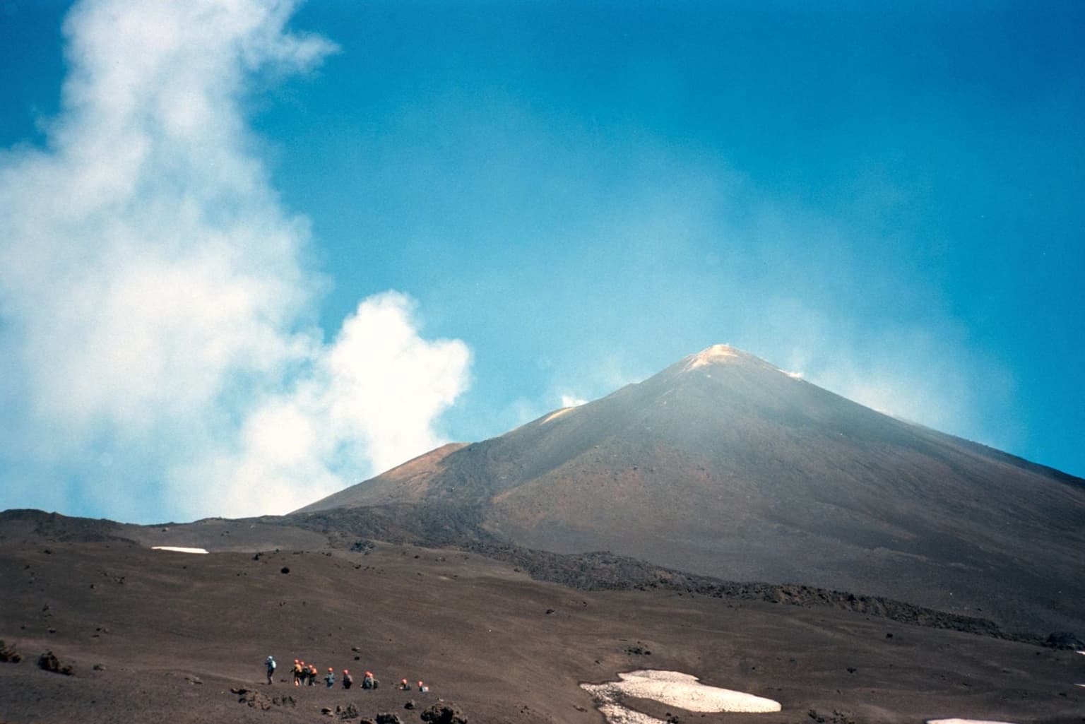 A line of hikers with backpacks climbs a bare black volcanic slope on Mount Etna, the mountain's smoking summit cone rising ahead under a clear blue sky.