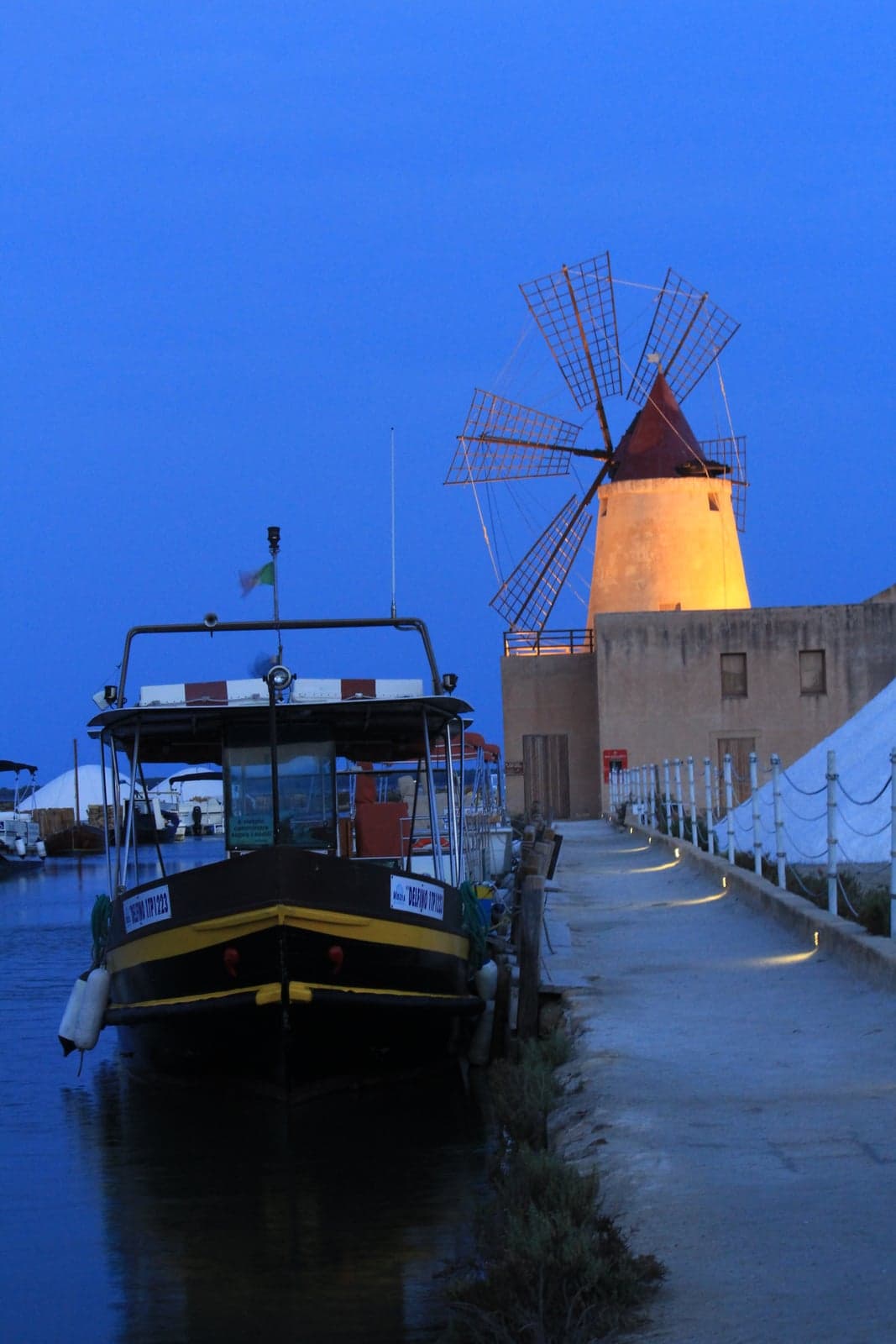 A passenger boat moored at the jetty at dusk, its canopy lit against a deep blue twilight sky, with the illuminated Mulino Infersa windmill and salt pans behind it