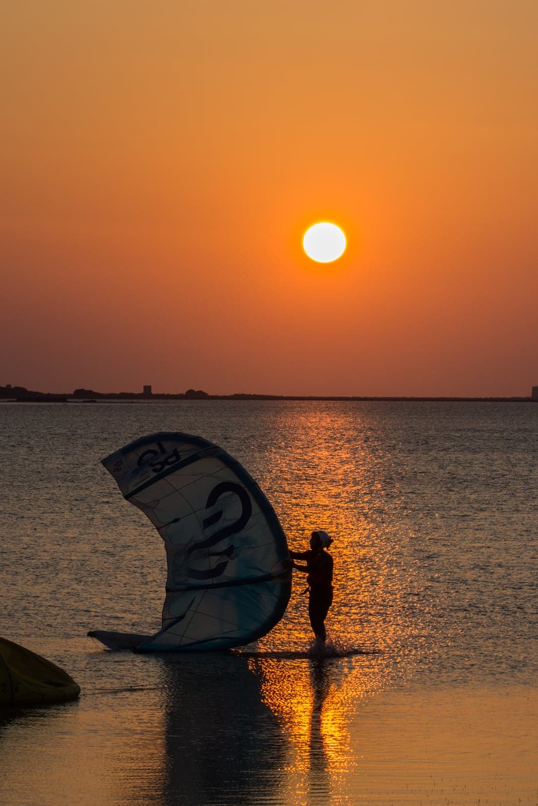 A kitesurfer standing shin-deep in the Stagnone at sunset, holding a large kite by its inflated edge as it rests half in the water, sunlight glittering across the shallow lagoon