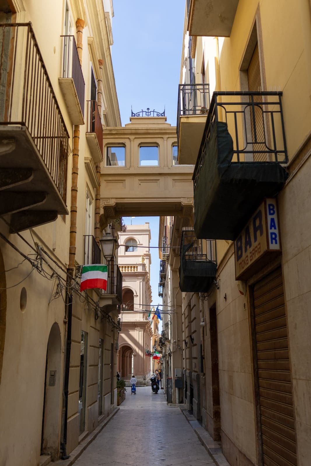 A narrow old-town alley in Marsala in the early morning, balconies and a covered skyway overhead, an Italian flag hanging from a lamp, a bar sign and a couple of people going about their day