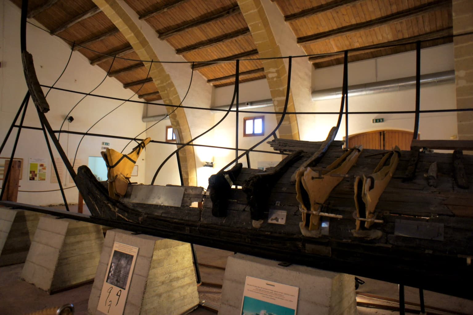 The full surviving length of the ancient Punic warship's hull, mounted on a metal armature that traces its original shape, displayed under the museum's vaulted wooden-beam ceiling