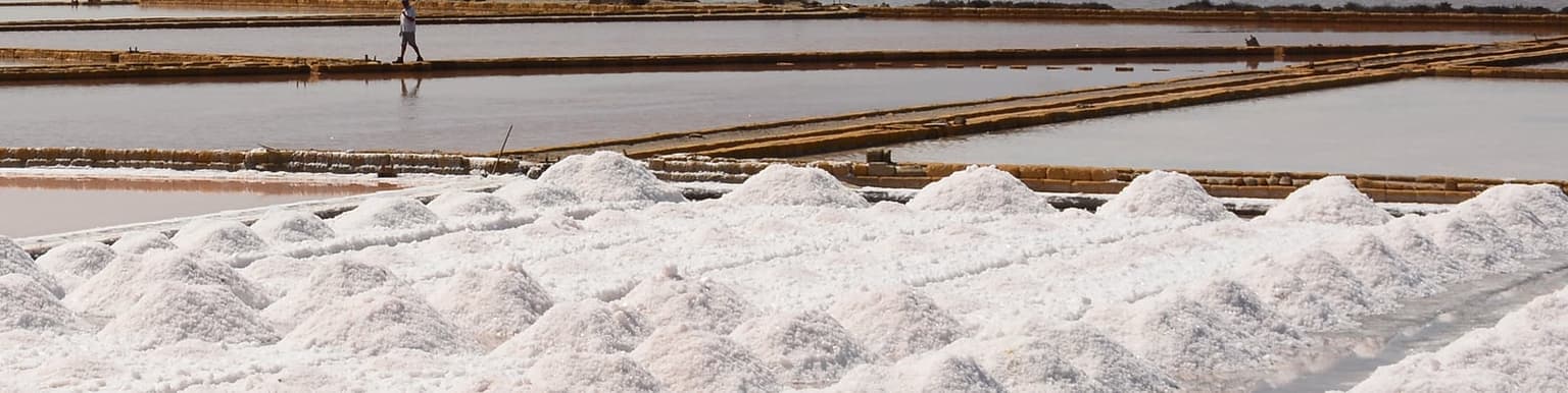 A close view of sharp white salt pyramids freshly piled at the water's edge, a worker walking the dyke behind them, the brine tinted pink
