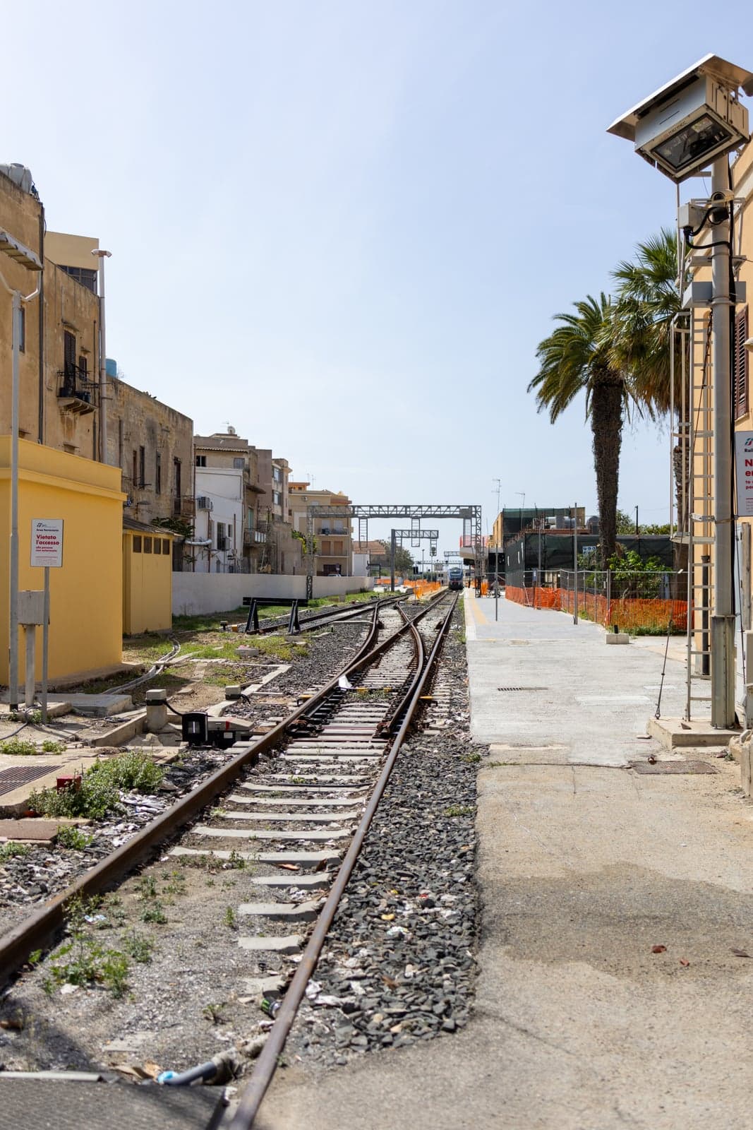Marsala's station platform and tracks running straight toward a distant regional train, a palm tree and old-town buildings lining the platform under a clear blue sky