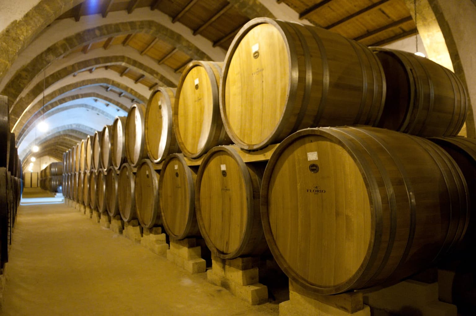 Rows of large oak barrels stamped FLORIO lining a long vaulted stone cellar, disappearing down the corridor under an arched brick ceiling