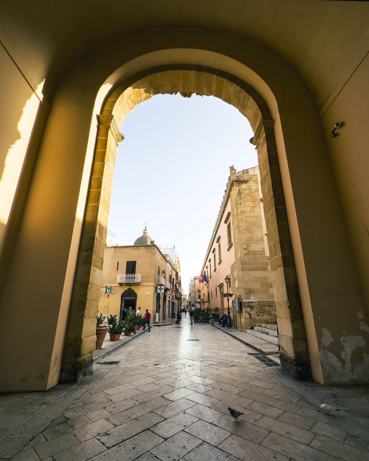A tall stone archway at golden hour framing the lit street of Marsala's old town beyond it, potted palms along the pavement and a church dome visible in the distance