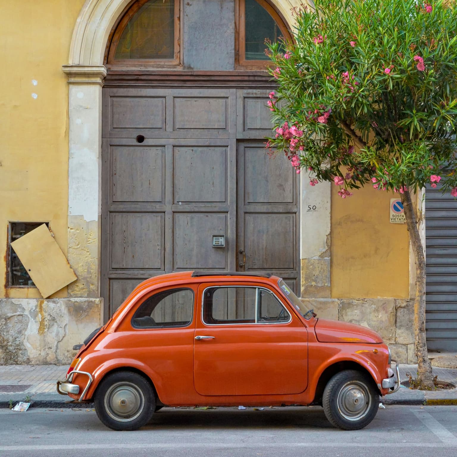 A restored orange Fiat 500 parked at the curb of an Italian street, in front of a weathered mustard-yellow building with tall wooden doors and a flowering oleander tree overhead
