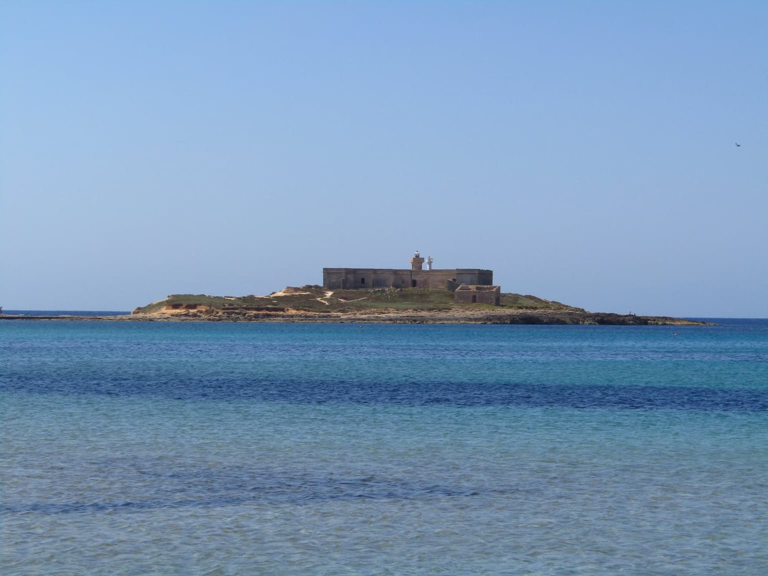 The lighthouse and fortress on Isola delle Correnti seen across turquoise water, the small island's low scrubby headland and stone buildings set against a clear blue sky