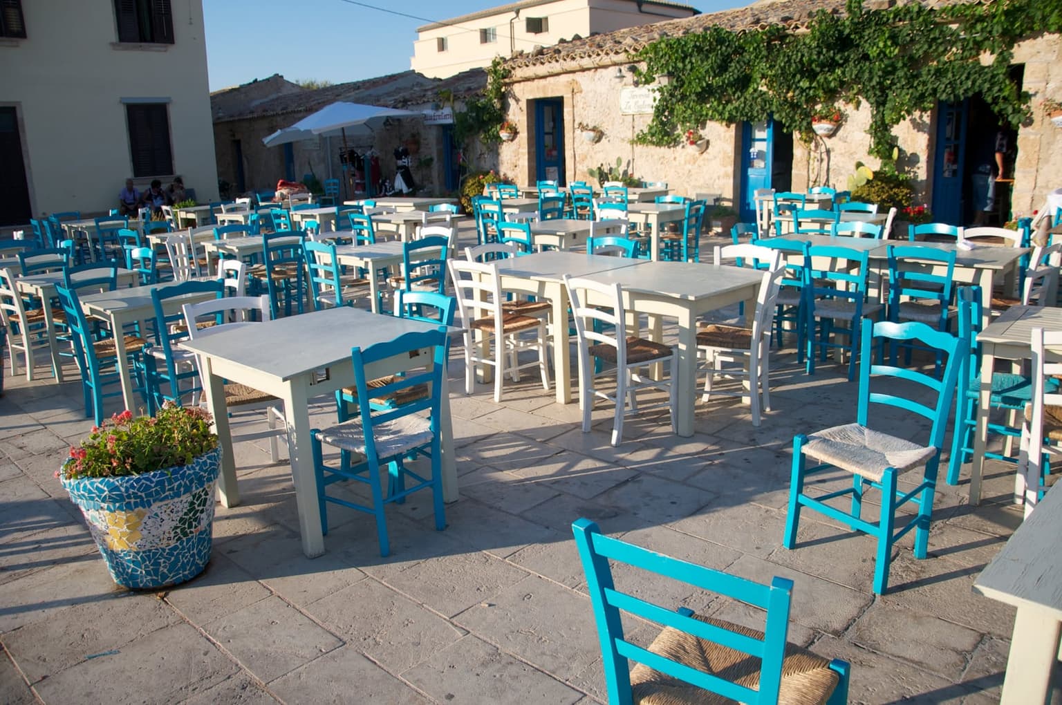 Rows of turquoise-blue wooden chairs and white tables set out on the stone paving of Marzamemi's piazza in late-afternoon sun, empty ahead of the dinner rush, with vine-draped stone buildings and open shopfronts behind