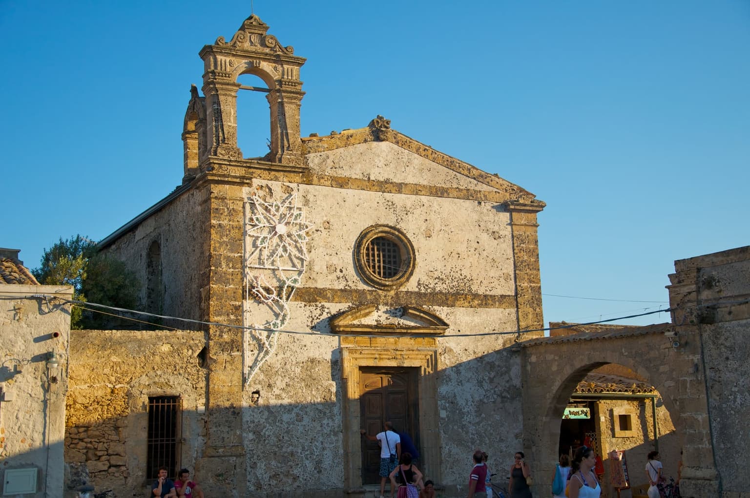 Weathered stone facade of a baroque church on Marzamemi's piazza, with its bell tower and round window catching late-afternoon sun, visitors gathered near the open wooden entrance door