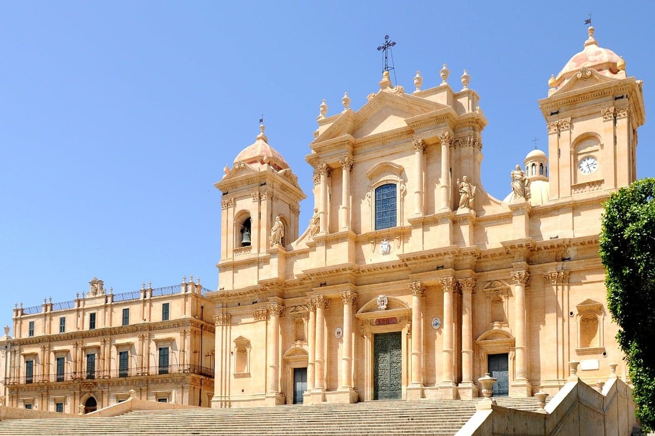 Noto Cathedral's honey-colored baroque facade rises behind its broad staircase, twin bell towers flanking a pedimented center with statues under a clear blue sky