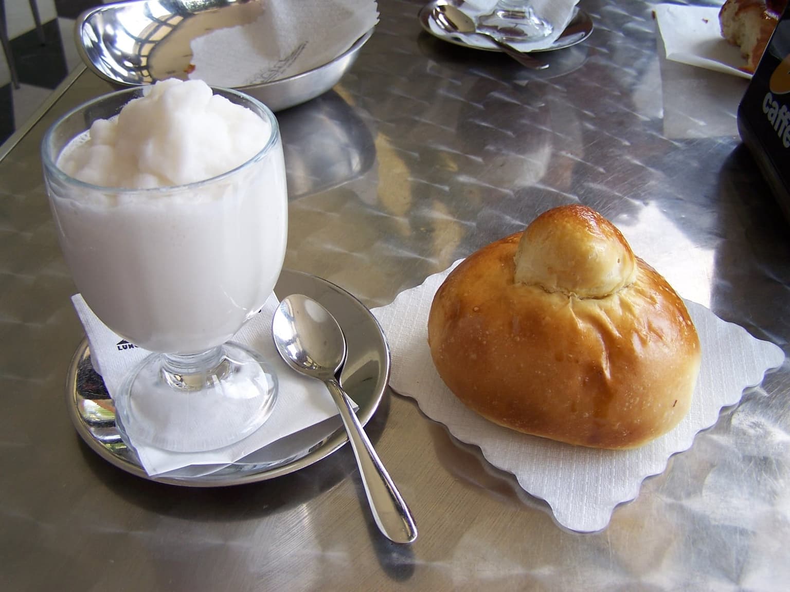 A footed glass of white almond granita with a spoon rests beside a glazed brioche col tuppo on a paper napkin, set on an ordinary stainless-steel café counter