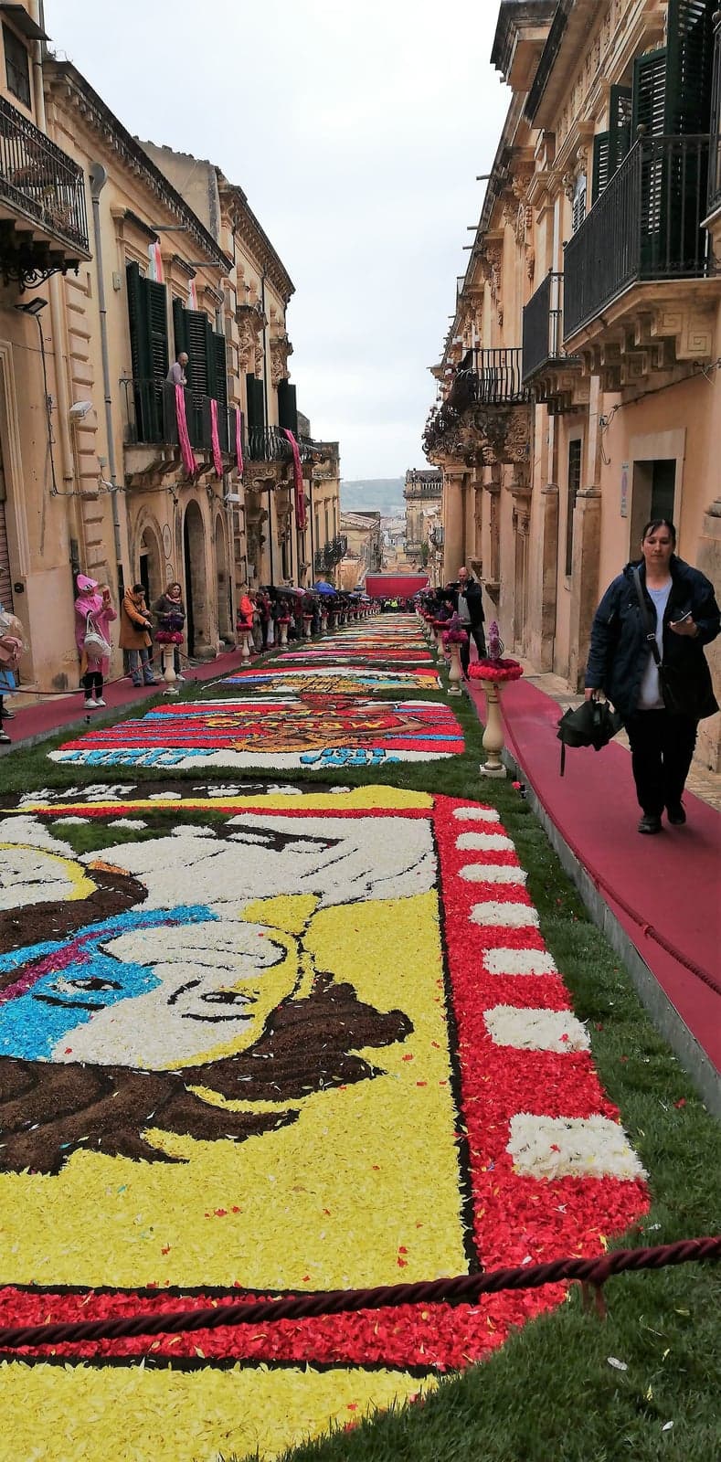 Elaborate flower-petal carpets in reds, yellows and blues fill a sloping street between balconied baroque palaces, as a woman walks along a red runway path beside the display under an overcast sky