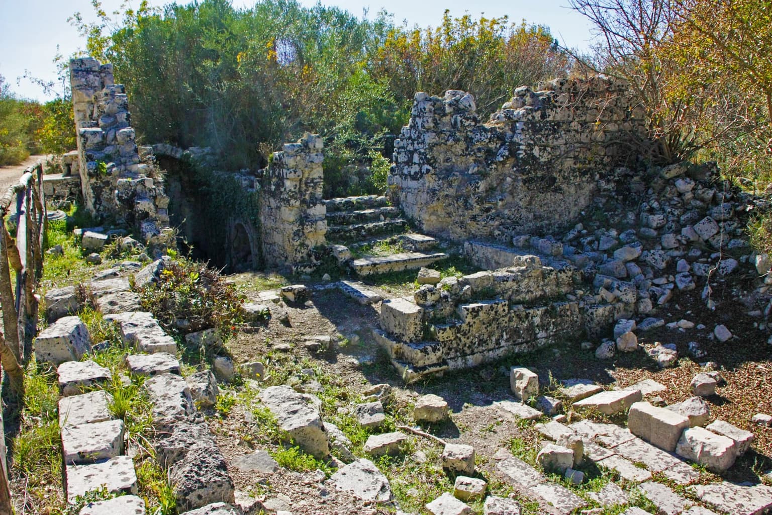 Overgrown limestone ruins at Noto Antica - broken walls, scattered cut blocks and a short stone stairway - sit half-buried in scrub and trees on the hillside, with no roof or standing structure left intact