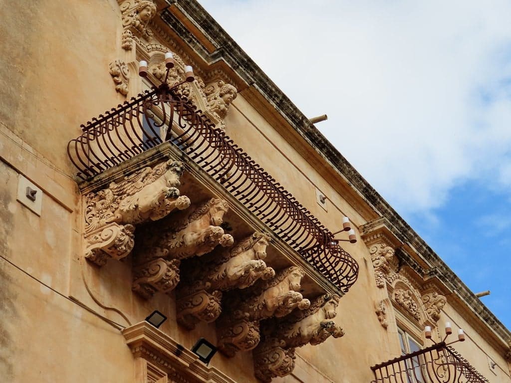 Carved stone corbels shaped like grotesque animal and mythic heads support a wrought-iron balcony on Palazzo Nicolaci's facade, photographed from directly below against a partly cloudy sky