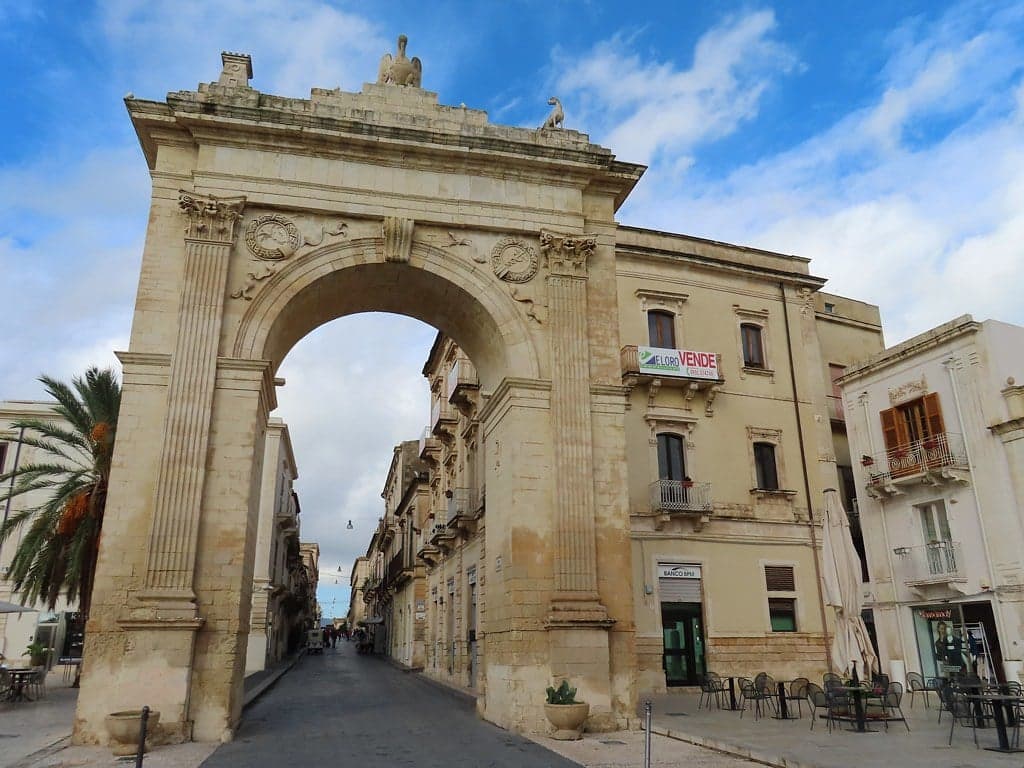 The Porta Reale, a triumphal stone arch topped with eagle and dog statues, frames the road leading into Noto's historic center, café tables and a palm tree beside it under a partly cloudy sky