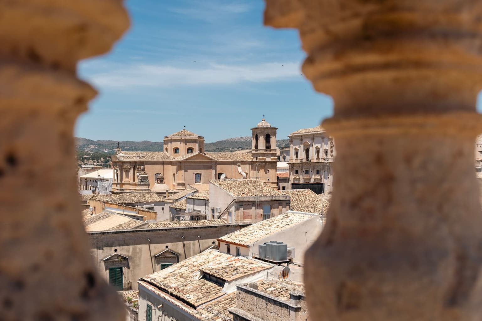 Looking through a gap in a stone parapet, terracotta rooftops spread out below toward a green-domed church tower and a bell tower, with hills in the distance