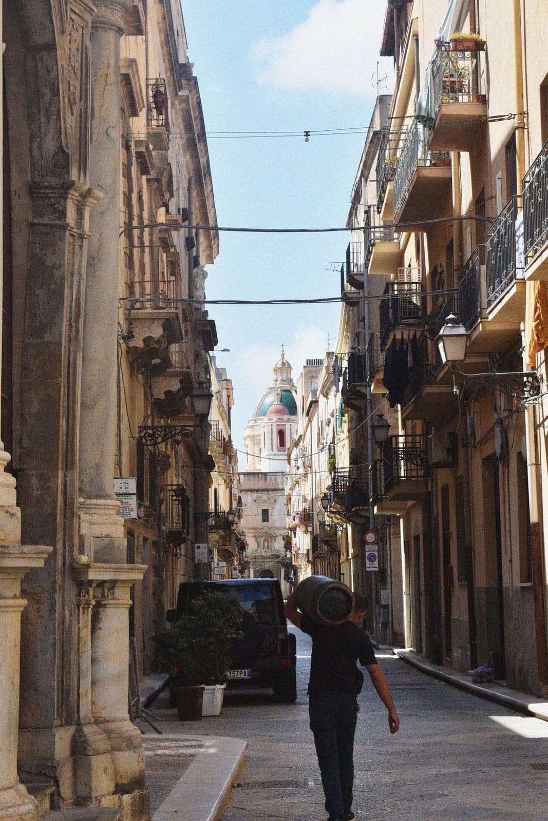 A narrow stone street lined with balconied buildings leads toward a distant green-domed church tower, where a man carrying a wooden barrel on his shoulder walks away from the camera past a parked van