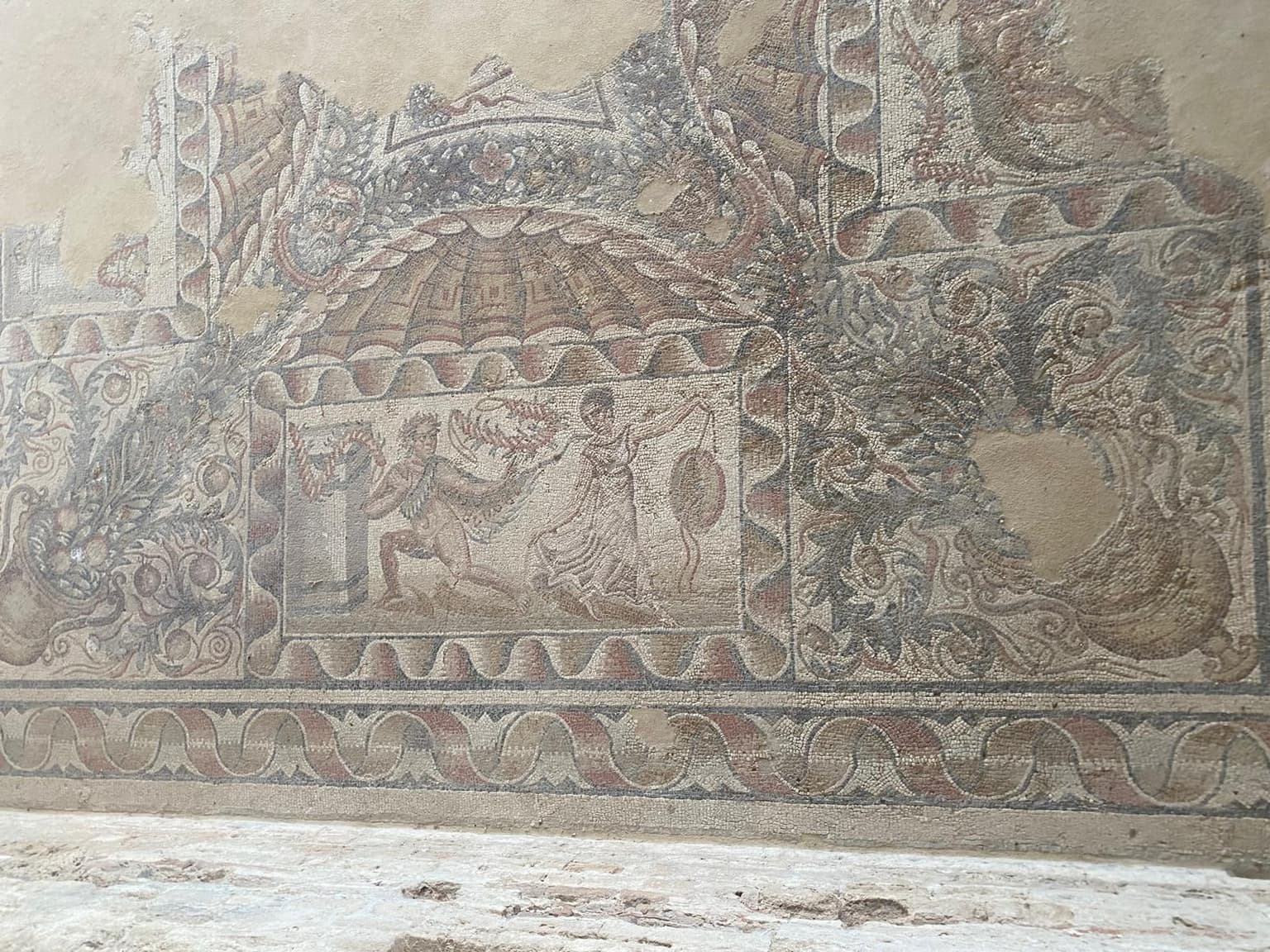 A weathered Roman floor mosaic at the Villa Romana del Tellaro shows a kneeling hunter reaching toward a draped figure beneath a scalloped canopy, framed by a wave-pattern border and acanthus scrollwork