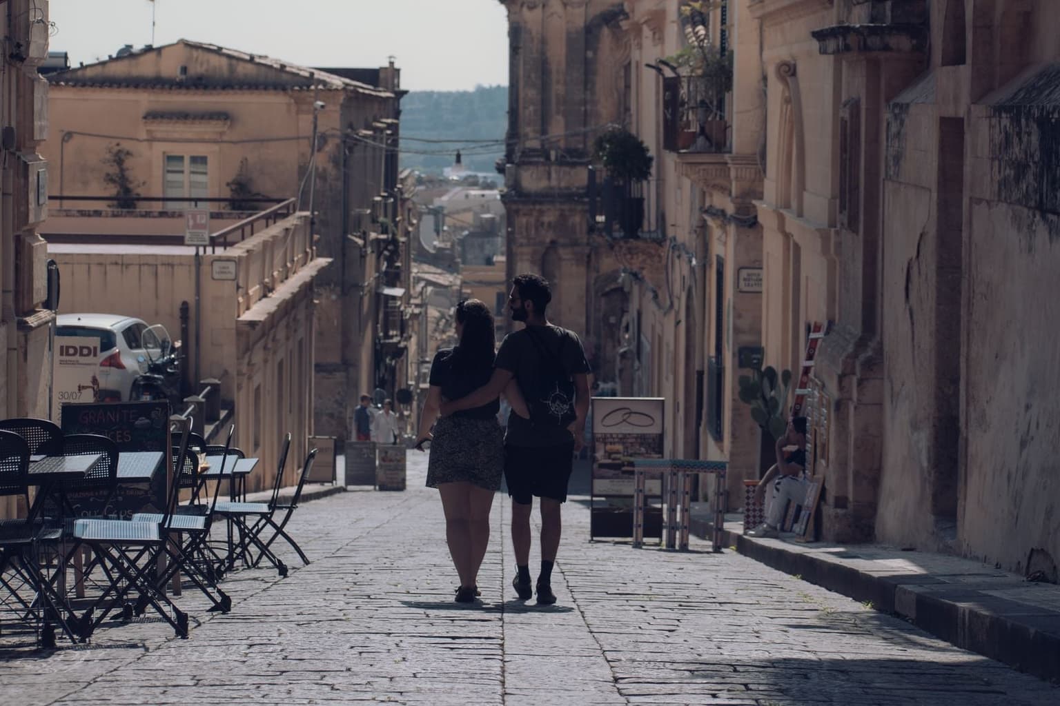 A couple walks away from the camera down a sunlit stone street lined with café tables and chairs, continuous baroque limestone facades running down both sides toward distant hills