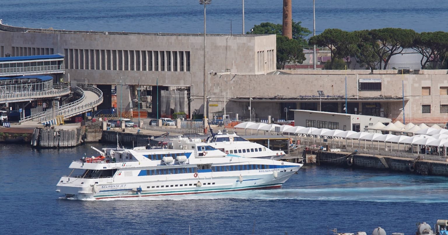 The Blu Jet fast boat Selinunte Jet crossing Messina harbor past the stone Stazione Marittima building, with a second fast boat moored at the covered quay behind it