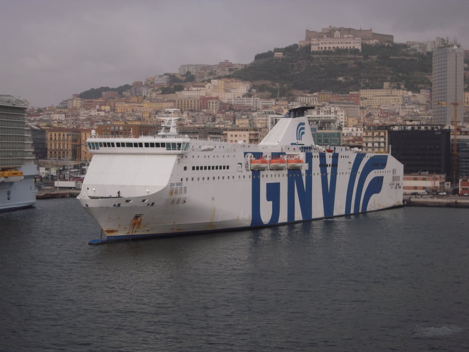 The white GNV Sirio ferry, its hull painted with large blue GNV letters, moored in the port of Naples under an overcast sky, with the hillside city and Castel Sant'Elmo rising behind