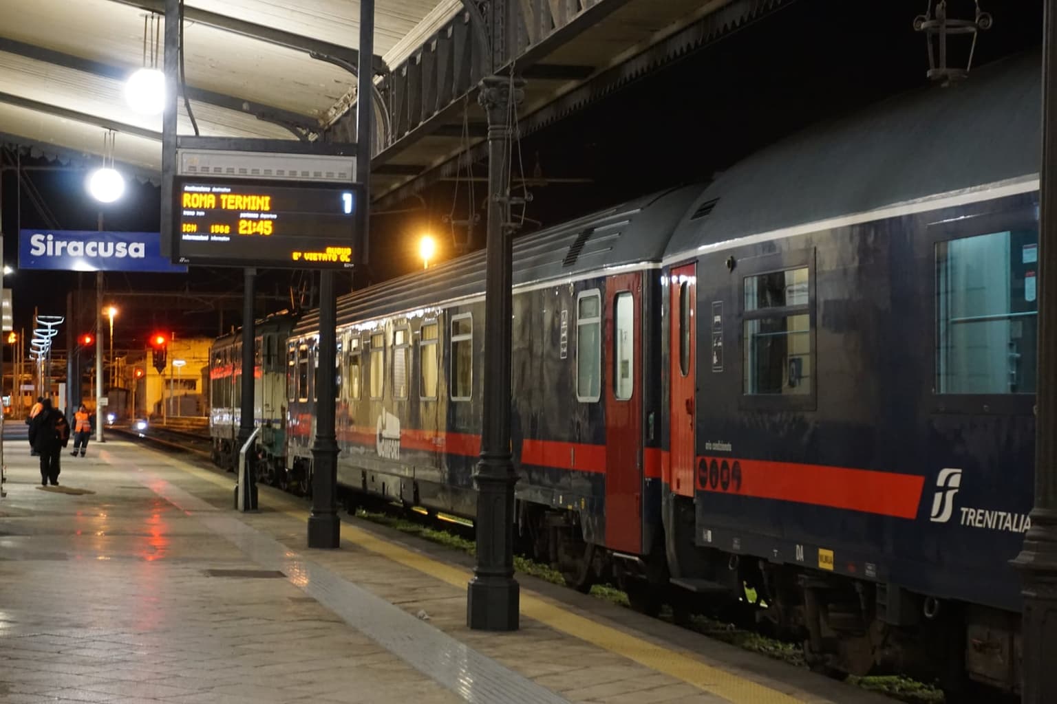 Dark blue Intercity Notte train cars at a wet platform in Siracusa station at night, under a Siracusa name board and a departure display for Roma Termini