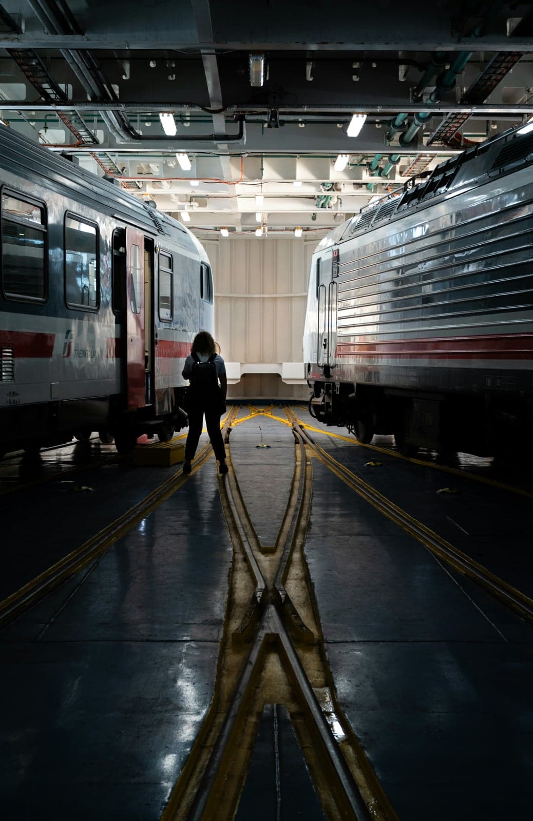 Inside a train ferry's hold, a Trenitalia Intercity passenger car and a silver locomotive stand on rails under a low steel ceiling of pipes and lights, with a passenger carrying a backpack between them