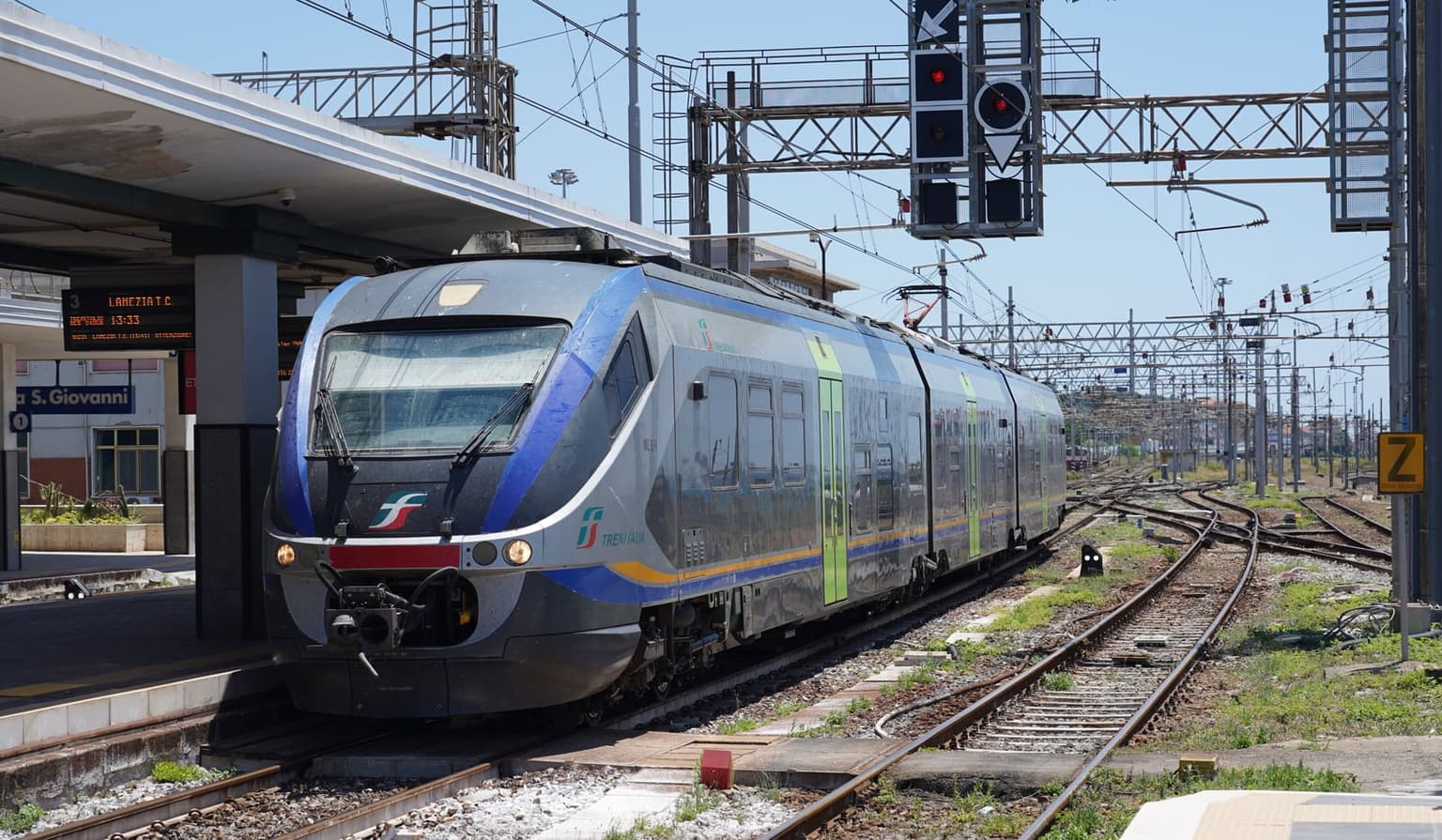 A blue and silver Trenitalia regional train at a platform in Villa San Giovanni station, with the blue station name board and a departure display for Lamezia Terme on the left