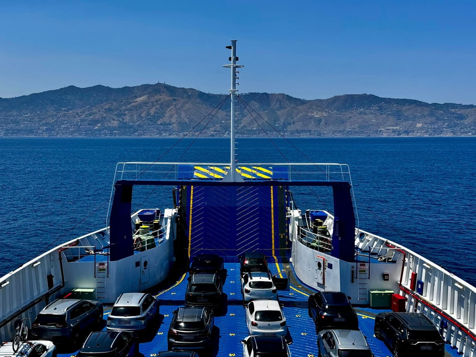 View from a car ferry's upper deck over rows of parked cars toward the open bow ramp, with deep blue water and a hilly coastline lined with towns ahead