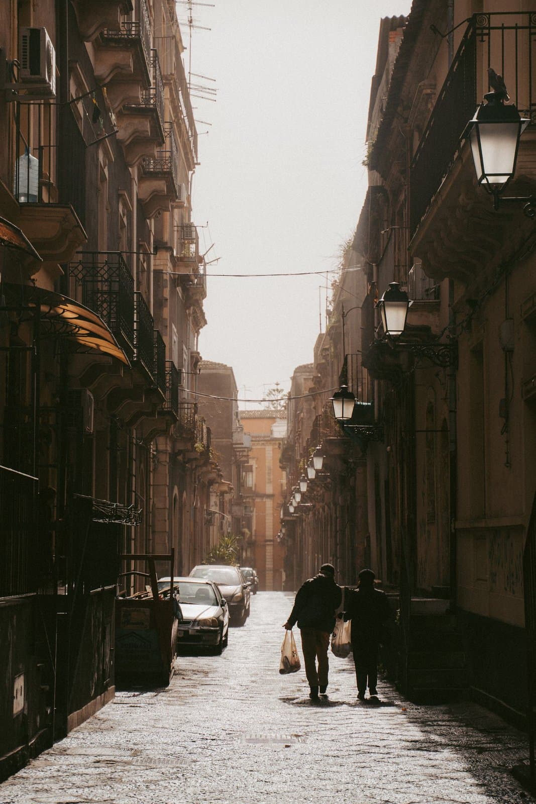 A narrow Catania street in hazy golden-hour light, wrought-iron balconies close overhead, cars parked tight along both sides and two locals walking home with grocery bags