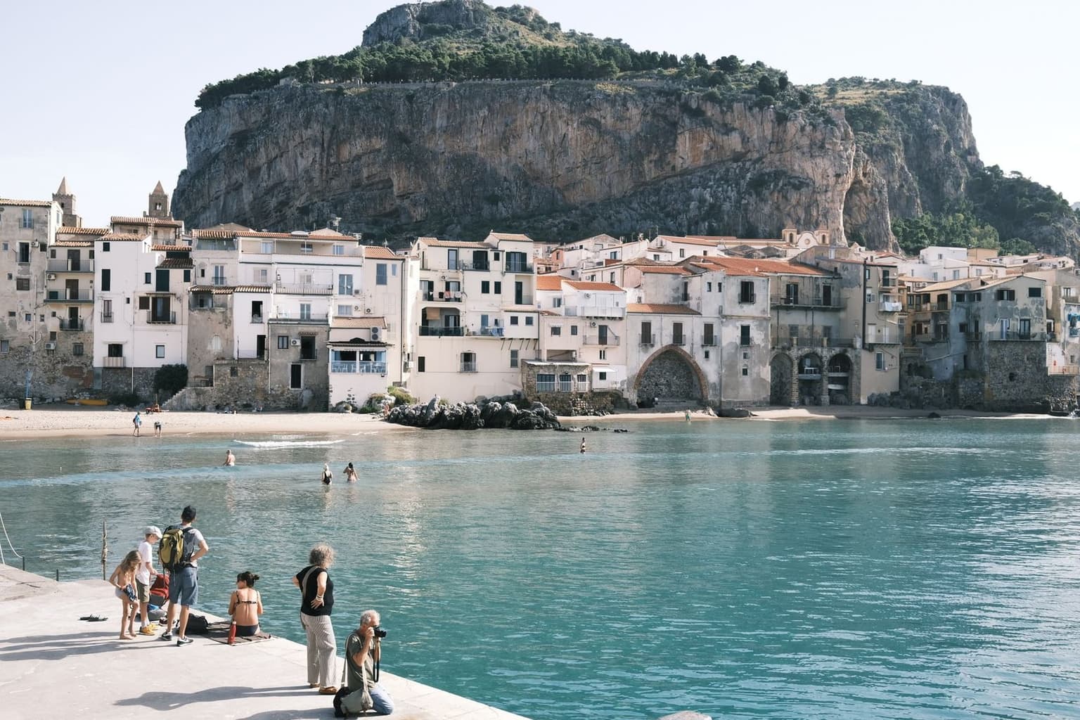 Cefalù's old town crowded against the sea below La Rocca, the sheer cliff rising behind the pastel waterfront houses, swimmers in the turquoise bay - the next base's view already opening up