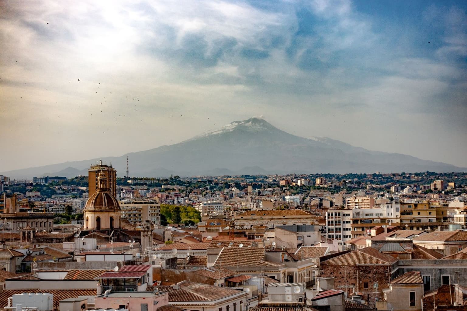 Mount Etna towering over Catania's terracotta rooftops and a church dome, a thin wisp of steam at the summit, evening light and clouds over the coastal plain