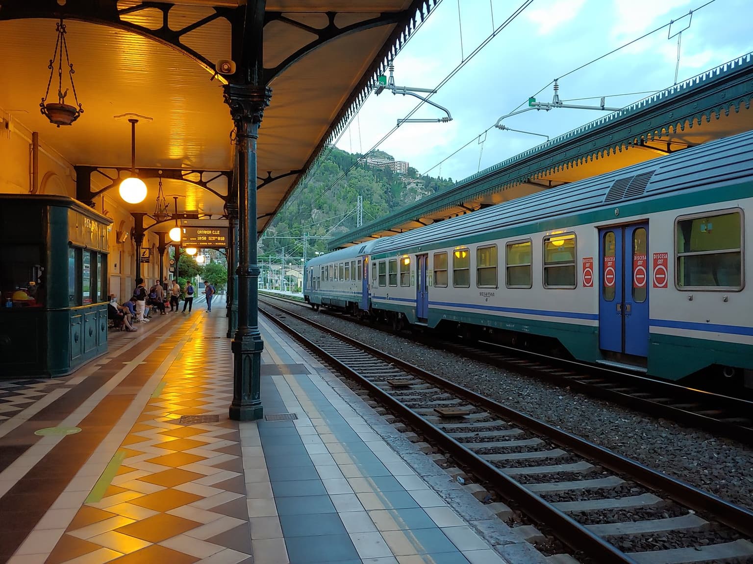 A regional train at Taormina-Giardini station in the evening, platform lights on, a wooded hillside rising beyond the tracks