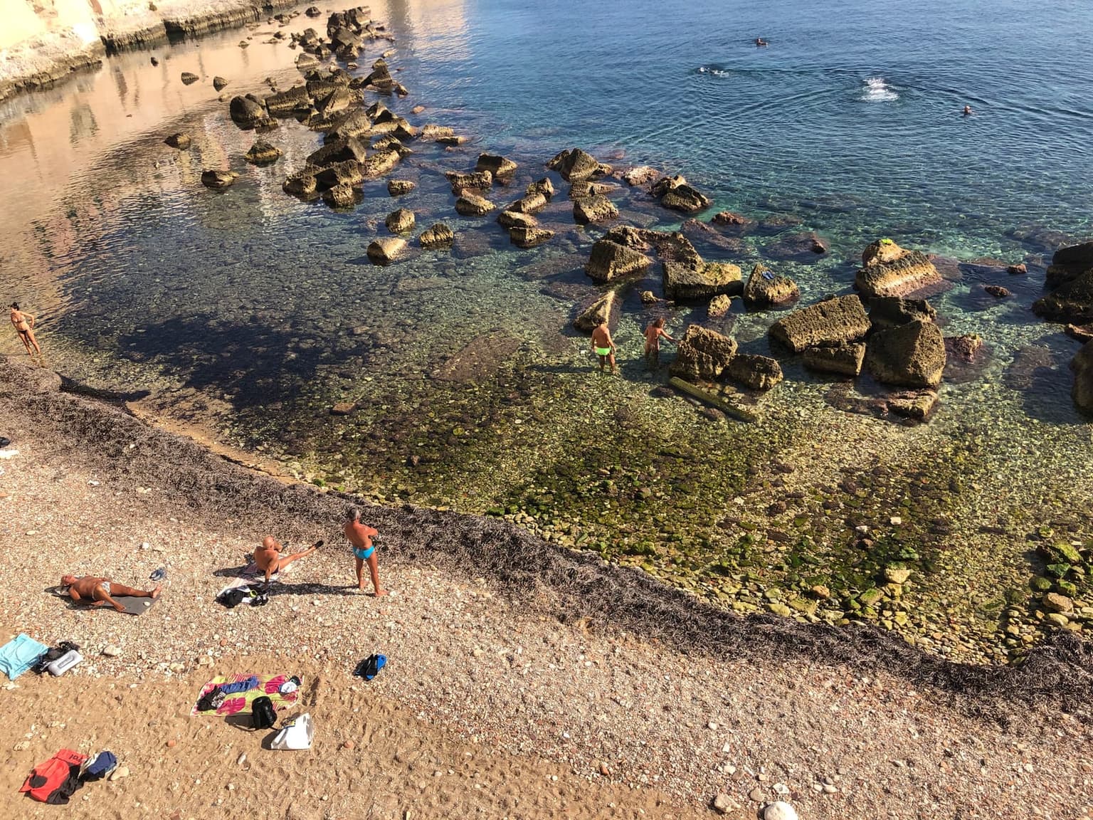 Cala Rossa, a small cove on Ortigia's edge where gravel is wedged between weathered rocks, people swimming in the clear water with the old town's stone wall behind