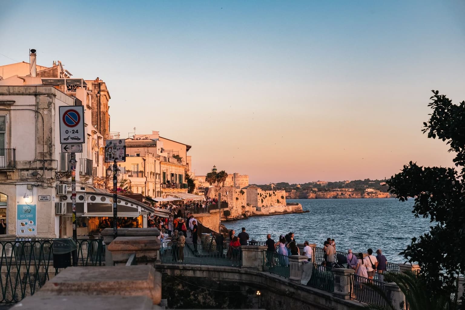 People walking Ortigia's seafront promenade at sunset, the old town's stone catching the last light
