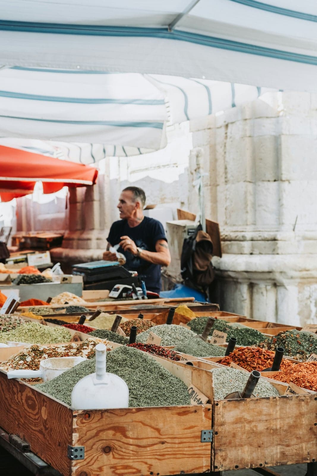 A vendor at Ortigia's market stalls, olives and spices in wooden crates, an ancient stone column base from the Temple of Apollo standing right behind the counter