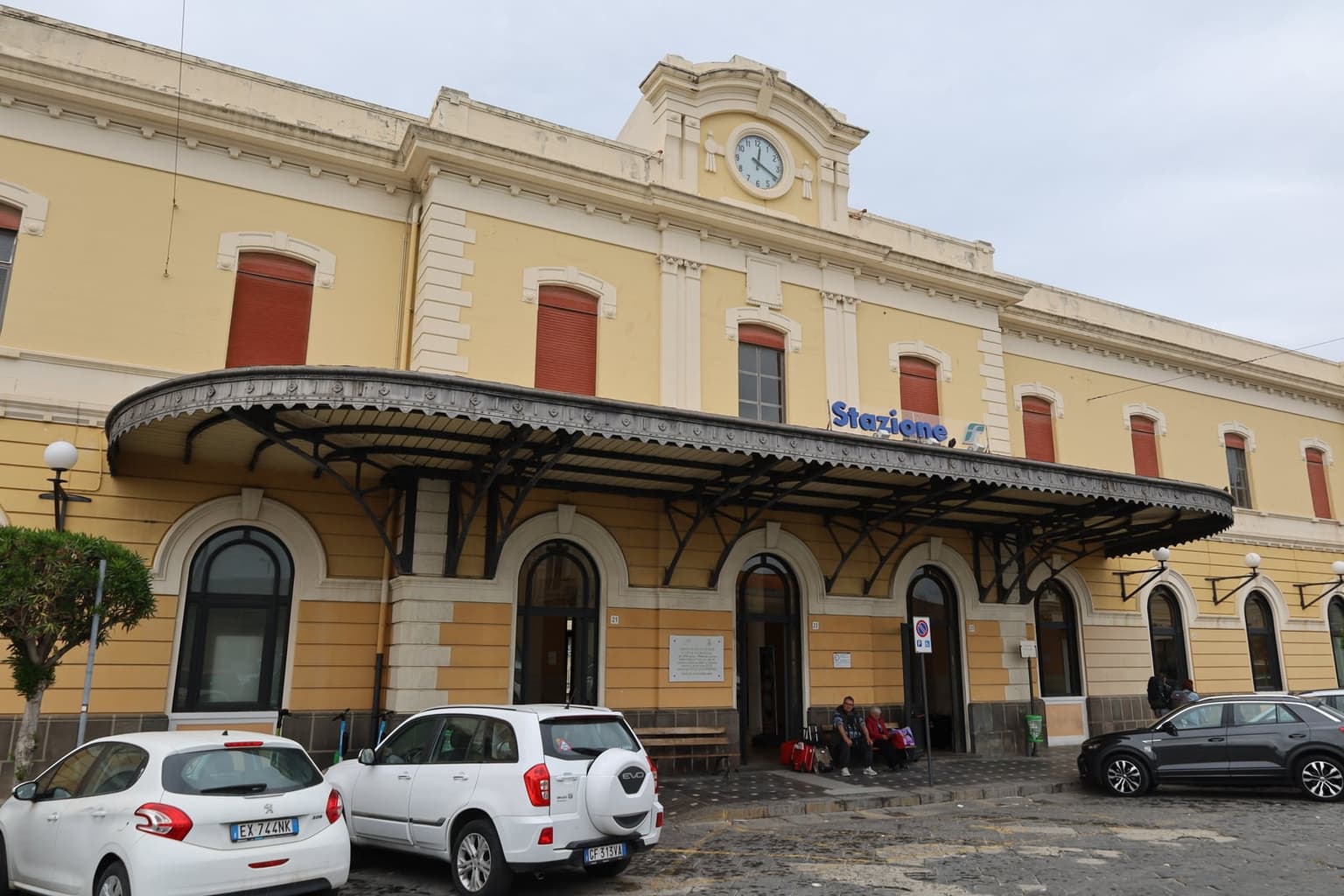 Siracusa's railway station, its yellow early-1900s façade and canopy, travelers with luggage waiting outside