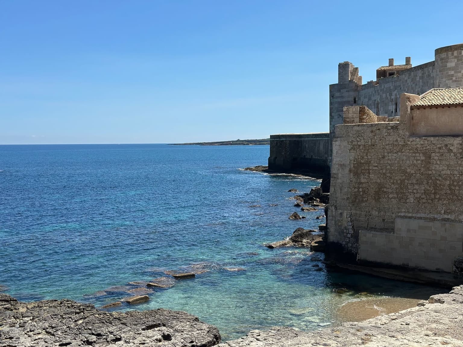Ortigia's fortified stone seafront wall dropping straight into clear turquoise water, the coastline curving away toward the horizon