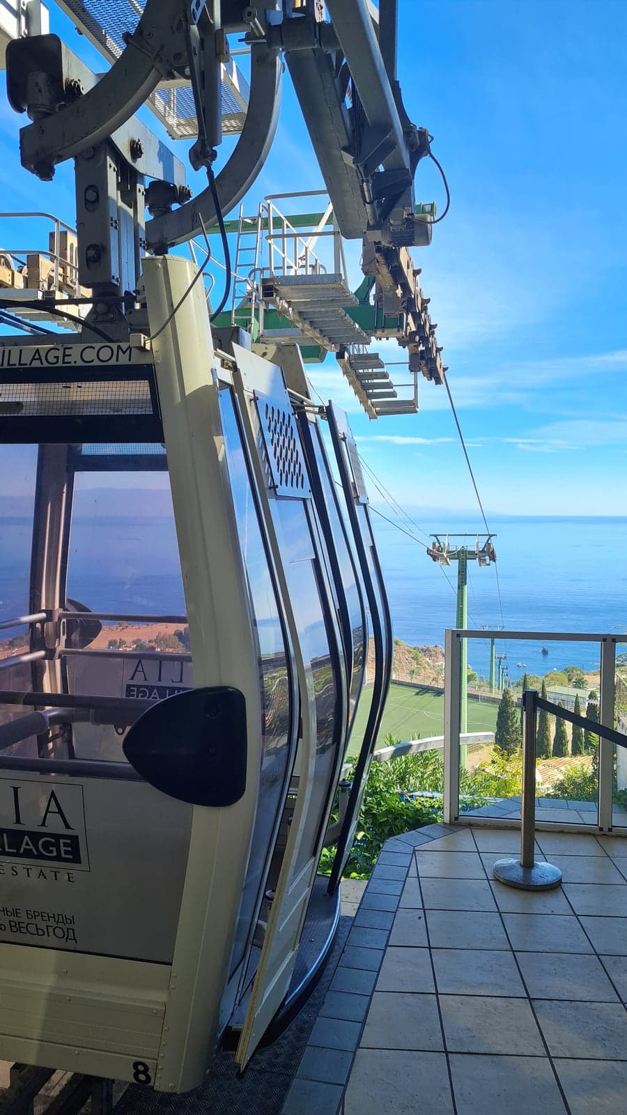 A cable-car cabin at the mountaintop station, the support cable and a lower tower visible against the sea and coastline below.