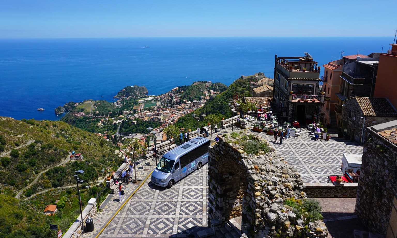View down from a hillside terrace above Taormina, the town's rooftops and the curving Ionian coast visible far below, with the rocky islet of Isola Bella in the distance.