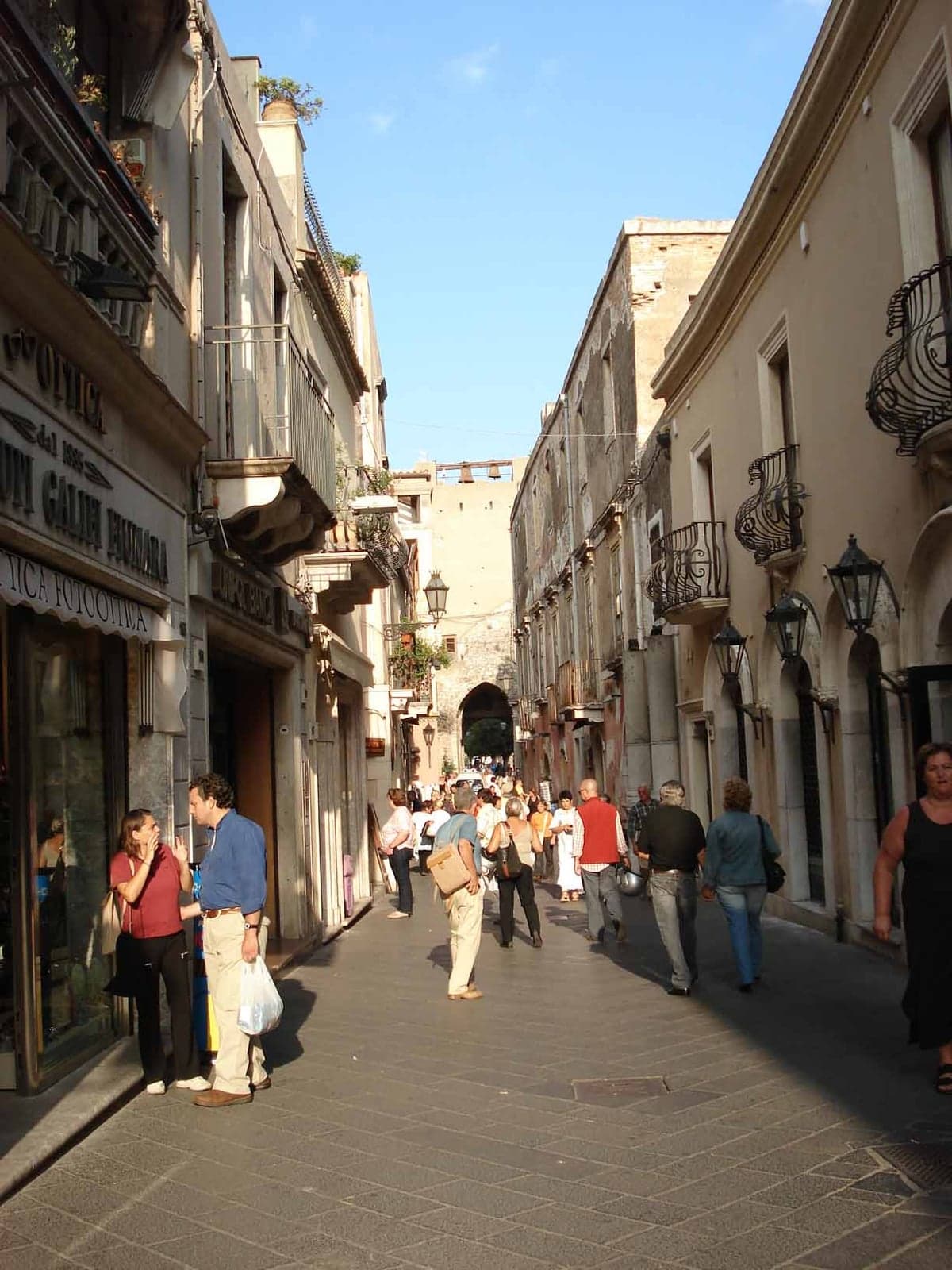 Corso Umberto packed with pedestrians between shopfronts and wrought-iron balconies, a stone archway visible at the far end of the street.